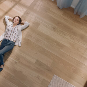 Young Woman is Lying on a Wooden Flooring in an Apartment. She's Dressed Casually. Cozy Living Room with Modern Minimalistic Interior and Wooden Parquet. Top View Camera Shot.